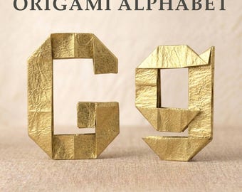 Könnte beinhalten: Der Großbuchstabe und Kleinbuchstabe G, gefertigt aus goldfarbenem Papier mittels Origami-Techniken. Das Papier weist eine ausgeprägte Knitterstruktur auf. Über den Buchstaben steht der Text ORIGAMI ALPHABET in einer dunkelgrauen serifenlosen Schrift.