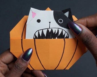 Könnte beinhalten: Ein orangefarbener Origami-Kürbis aus Papier mit handgezeichneten schwarzen vertikalen Linien. Im Kürbis befindet sich ein weiß-schwarzes Origami-Katzengesicht mit rosa Innenohren und einem breiten, offenen Mund mit spitzen, schwarzen Zähnen.