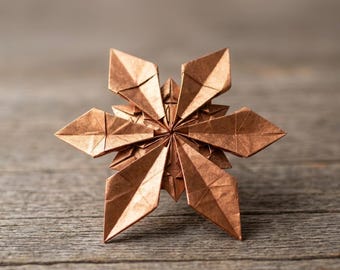 Könnte beinhalten: Ein kupferfarbener Origami-Stern mit sechs spitzen Blütenblättern, die in einem symmetrischen, geschichteten geometrischen Muster angeordnet sind. Die Papierskulptur weist scharfe, präzise Falten und einen metallischen Glanz auf, der eine komplexe, mehrdimensionale Blumenform erzeugt.