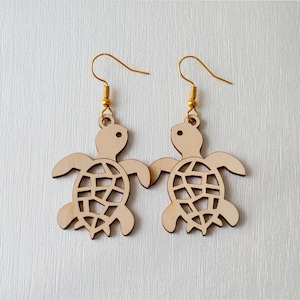 Op de afbeelding: Een paar houten oorbellen in de vorm van een schildpad. Elke oorbel heeft een lichtbruine schildpadvorm met een uitgesneden schildontwerp. De oorbellen hangen aan goudkleurige oorhaakjes. De schildpadden hebben kleine, ronde ogen.