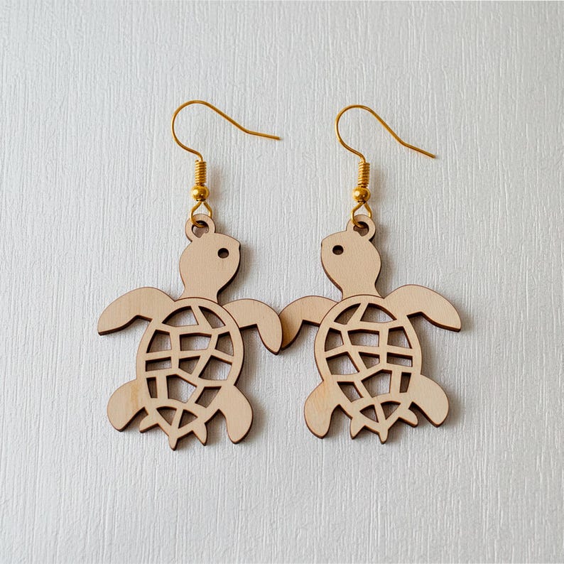 Op de afbeelding: Een paar houten oorbellen in de vorm van een schildpad. Elke oorbel heeft een lichtbruine schildpadvorm met een uitgesneden schildontwerp. De oorbellen hangen aan goudkleurige oorhaakjes. De schildpadden hebben kleine, ronde ogen.