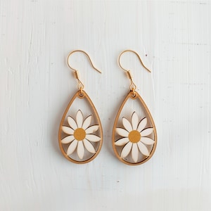 Könnte beinhalten: Ein Paar tropfenförmige Holzohrringe mit Gänseblümchen-Design. Die Ohrringe haben einen hellbraunen Holzrahmen mit einer weißen und gelben Gänseblümchenblüte in der Mitte. Die Ohrhaken sind goldfarben.