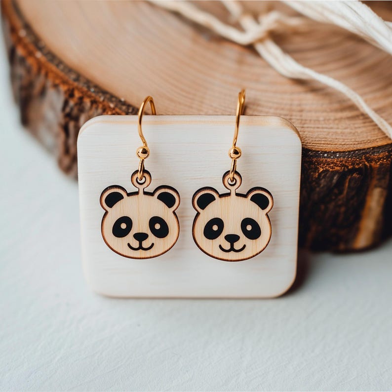 Op de afbeelding: Een paar panda-oorbellen. Elke oorbel heeft een lichtgekleurd houten pandagezicht met zwarte ogen, neus en omtreklijnen. De oorbellen hangen aan goudkleurige haken. De oorbellen worden gepresenteerd op een klein, vierkant, lichtgekleurd houten blok.