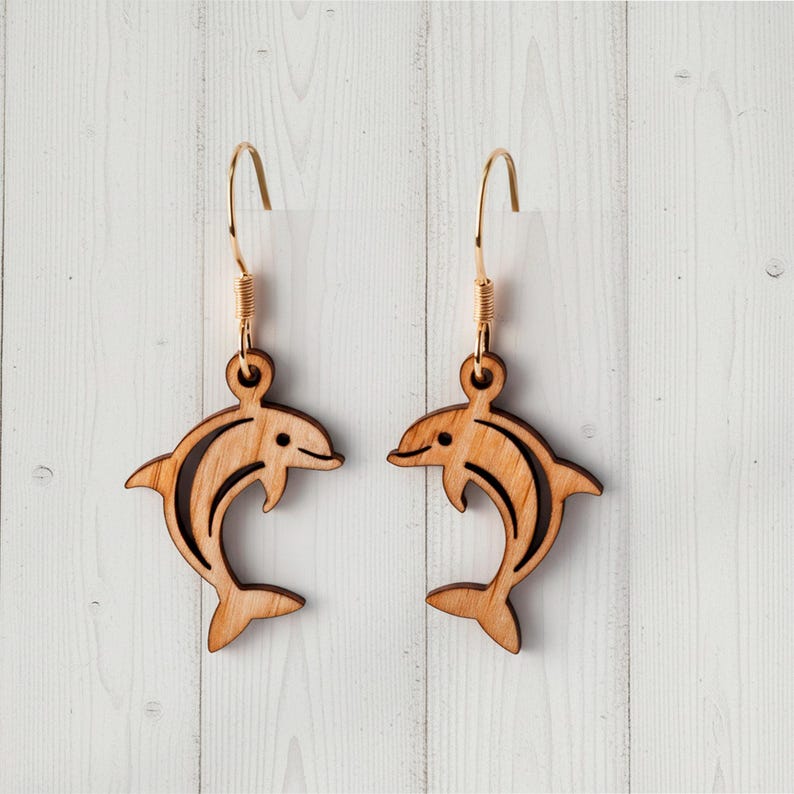Op de afbeelding: Een paar houten dolfijn oorbellen. Elke oorbel heeft een dolfijn silhouet met ge&euml;tste details en een goudkleurige haak. De dolfijnen zijn lichtbruin en hebben een gladde afwerking. De oorbellen worden gepresenteerd tegen een witte houten achtergrond.
