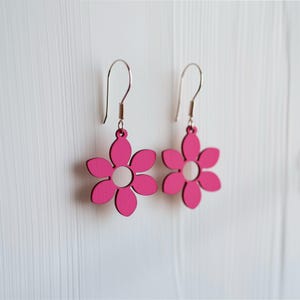 Op de afbeelding: Een paar felroze bloemvormige oorbellen met zilverkleurige haken. Elke oorbel heeft een bloemontwerp met vijf bloemblaadjes en een wit midden. De oorbellen hangen tegen een witte achtergrond.