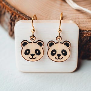 May include: A pair of wooden panda bear earrings with gold-colored hooks. The earrings feature a light brown panda face with black eyes, ears, and a nose. The earrings are displayed on a small, square, light-colored wooden block.