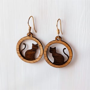 Op de afbeelding: Een paar houten kattenoorbellen. Elke oorbel heeft een donkerbruin katten silhouet in een lichtbruin cirkelvormig frame. De oorbellen hebben goudkleurige oorhaken.