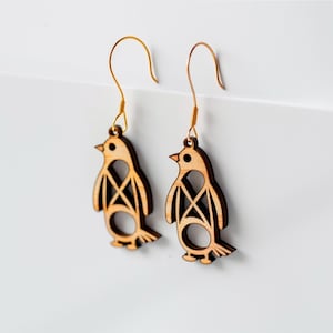 Op de afbeelding: Een paar houten pingu&iuml;n oorbellen met goudkleurige haken. De oorbellen zijn lichtbruin met een uitgesneden ontwerp op het lichaam. De snavel en poten van de pingu&iuml;n zijn zichtbaar. De oorbellen hangen tegen een witte achtergrond.