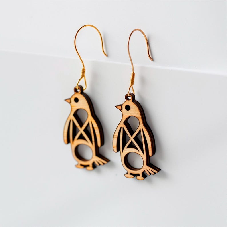Op de afbeelding: Een paar houten pingu&iuml;n oorbellen met goudkleurige haken. De oorbellen zijn lichtbruin met een uitgesneden ontwerp op het lichaam. De snavel en poten van de pingu&iuml;n zijn zichtbaar. De oorbellen hangen tegen een witte achtergrond.