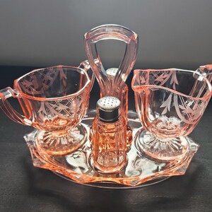 May include: A vintage pink glass condiment set. The set includes a sugar bowl, creamer, salt and pepper shakers, and a serving tray. The pieces have etched floral designs. The tray has a handle for easy carrying. A classic addition to any table setting.