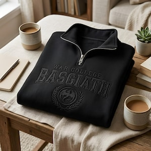 Puede incluir: Sudadera negra con cuello con cremallera. La sudadera presenta el texto "WAR COLLEGE BASGIATH" en relieve, junto con un emblema de drag&oacute;n. La sudadera est&aacute; doblada sobre una mesa de madera, con tazas de caf&eacute; y un cuaderno cerca.