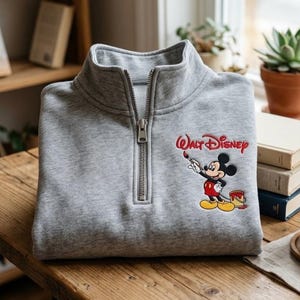 Puede incluir: Una sudadera gris con cremallera de cuarto de cuello con un gráfico de Mickey Mouse y el texto "Walt Disney". El gráfico muestra a Mickey Mouse sosteniendo un pincel y un cubo de pintura. La sudadera está doblada sobre una mesa de madera.