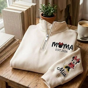 May include: Cream-colored quarter-zip sweatshirt with the word "MOMA" in black, a Minnie Mouse head, and "EST.2026". The sleeve features a Minnie Mouse graphic and the name "Mia". The sweatshirt is on a wooden table.