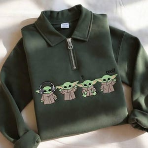 May include: Dark green sweatshirt with a collared neckline and a quarter-zip closure. The front features an embroidered design of four Grogu characters, with the word "cool!" printed on the right side.
