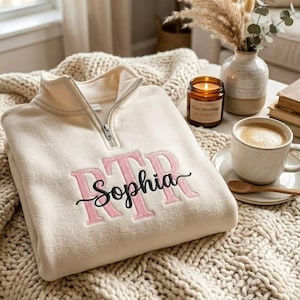 May include: Cream-colored quarter-zip sweatshirt with the name "Sophia" embroidered in pink and black. The sweatshirt is folded on a textured blanket, next to a cup of coffee, a candle, and a vase of dried flowers.