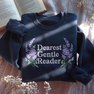 Puede incluir: Una sudadera azul marino con la frase bordada "Dearest Gentle Reader" rodeada de ramitas de lavanda. Un libro abierto es visible en el fondo. La sudadera está doblada.