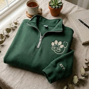 May include: A dark green quarter-zip sweatshirt with a white embroidered paw print and stethoscope design that says "Vet Tech". The sleeve cuff has the initials "RVT" embroidered in white.