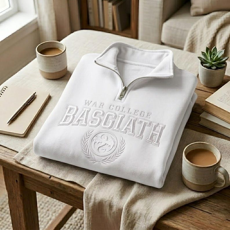 Puede incluir: Una sudadera blanca doblada con cuello con cremallera. La sudadera presenta el texto bordado "WAR COLLEGE BASGIATH" con un dise&ntilde;o de escudo. Tambi&eacute;n se ven dos tazas de caf&eacute;, una planta suculenta y un bloc de notas.