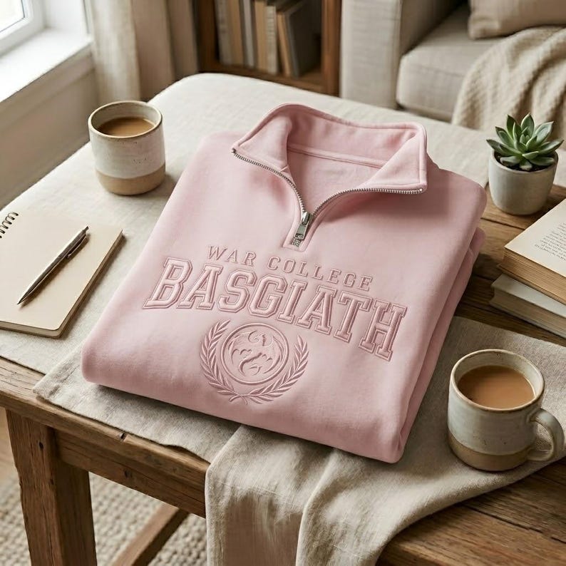 Puede incluir: Sudadera rosa claro con cuello con cremallera y el texto bordado "WAR COLLEGE BASGIATH". El dise&ntilde;o incluye un escudo con una corona de laurel. La sudadera est&aacute; doblada sobre una mesa de madera, junto a un cuaderno, un bol&iacute;grafo y dos tazas de caf&eacute;.