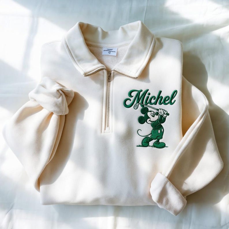 May include: Cream-colored sweatshirt with a quarter-zip collar and long sleeves. Features a green embroidered graphic of a cartoon mouse playing golf, with the name "Michel" above. The sweatshirt is folded neatly.