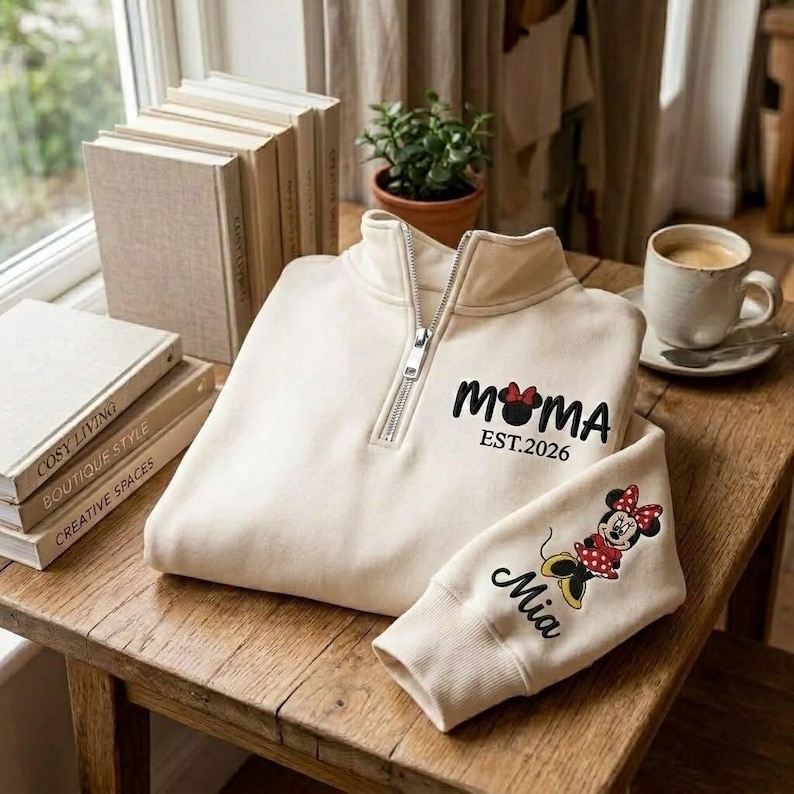 May include: Cream-colored quarter-zip sweatshirt with the word "MOMA" in black, a Minnie Mouse head, and "EST.2026". The sleeve features a Minnie Mouse graphic and the name "Mia". The sweatshirt is on a wooden table.