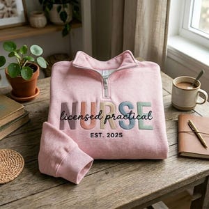 May include: A pale pink sweatshirt with a quarter-zip collar. The word "NURSE" is embroidered in various colours, with "licensed practical" and "EST. 2025" below. The sweatshirt is folded on a wooden table.