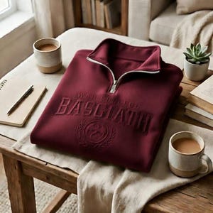 Puede incluir: Una sudadera burdeos con cremallera de un cuarto con el texto "WAR COLLEGE BASCIATH" bordado en el pecho. La sudadera tiene una cremallera plateada y un peque&ntilde;o emblema debajo del texto. La imagen est&aacute; sobre una mesa de madera con tazas de caf&eacute; y una suculenta.