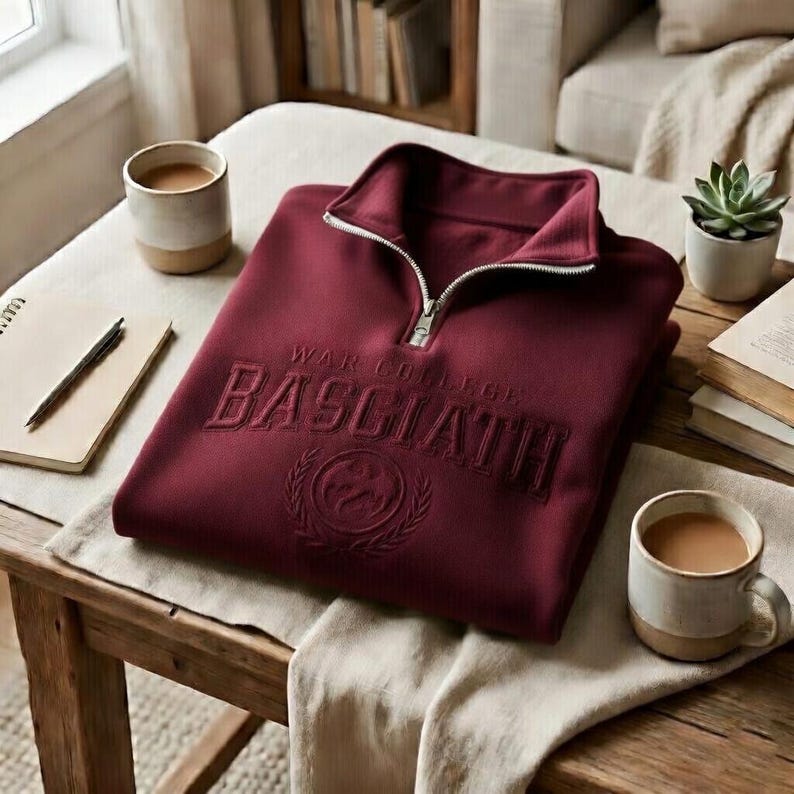 Puede incluir: Una sudadera burdeos con cremallera de un cuarto con el texto "WAR COLLEGE BASCIATH" bordado en el pecho. La sudadera tiene una cremallera plateada y un peque&ntilde;o emblema debajo del texto. La imagen est&aacute; sobre una mesa de madera con tazas de caf&eacute; y una suculenta.