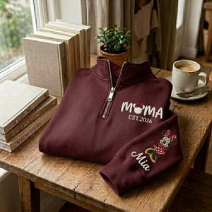 May include: Burgundy quarter-zip sweatshirt with "MAMA" and a Mickey Mouse head design, "EST. 2026" text, and "Mia" embroidered on the sleeve. The sweatshirt is on a wooden table with books and a cup of coffee.
