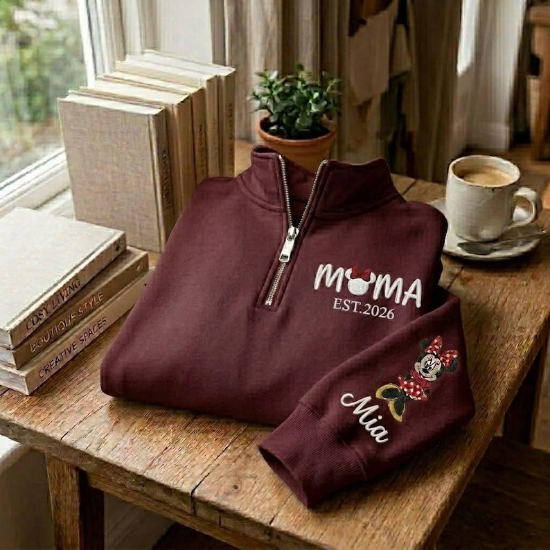 May include: Burgundy quarter-zip sweatshirt with "MAMA" and a Mickey Mouse head design, "EST. 2026" text, and "Mia" embroidered on the sleeve. The sweatshirt is on a wooden table with books and a cup of coffee.