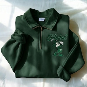 May include: Dark green sweatshirt with a collared neckline and a quarter-zip closure. Features an embroidered Mickey Mouse golfing graphic and the name "Michel" in script. The sleeves are cuffed.