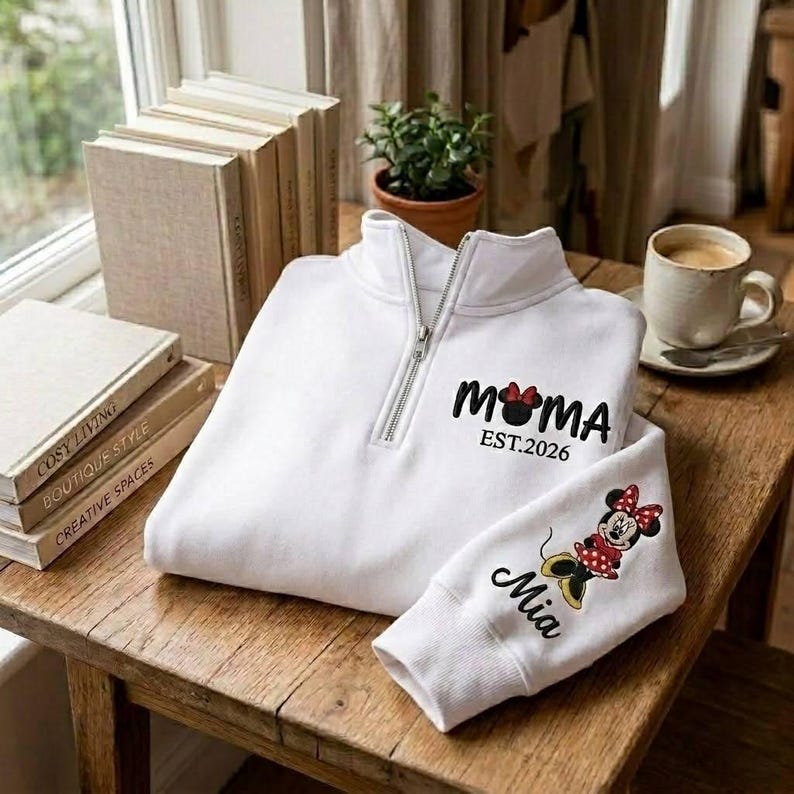 May include: A white quarter-zip sweatshirt with the word "MOMA" and a Minnie Mouse graphic, along with "EST.2026" printed on the front. The sleeve features a Minnie Mouse graphic and the name "Mia". The sweatshirt is folded on a wooden table.