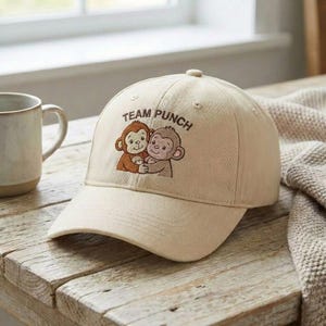 Peut inclure: Casquette de baseball beige avec l'inscription "TEAM PUNCH" et une image brodée de deux singes de dessin animé qui s'embrassent. La casquette est posée sur une table en bois, à côté d'une tasse et d'une couverture en tricot.