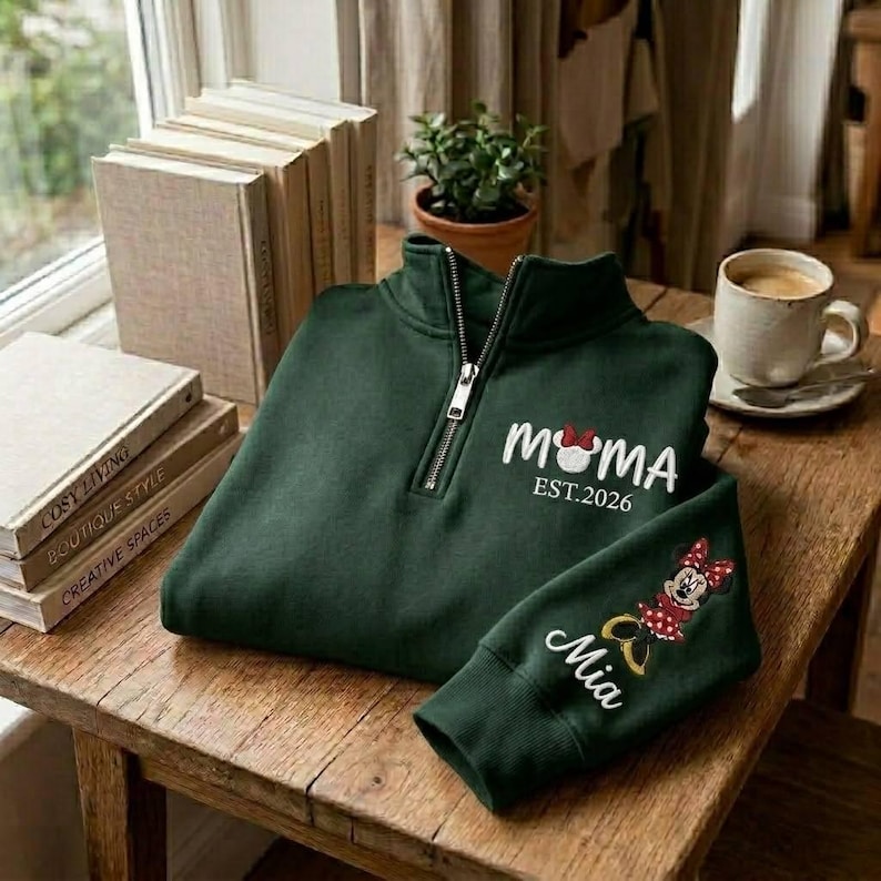 May include: A dark green quarter-zip sweatshirt with the word "MOMA" and a Minnie Mouse graphic, with "EST.2026" printed below. The sleeve features a Minnie Mouse graphic and the name "Mia". The sweatshirt is on a wooden table.