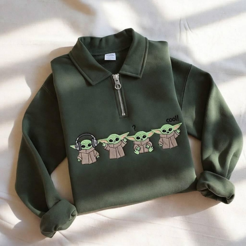 May include: Dark green sweatshirt with a collared neckline and a quarter-zip closure. The front features an embroidered design of four Grogu characters, with the word "cool!" printed on the right side.