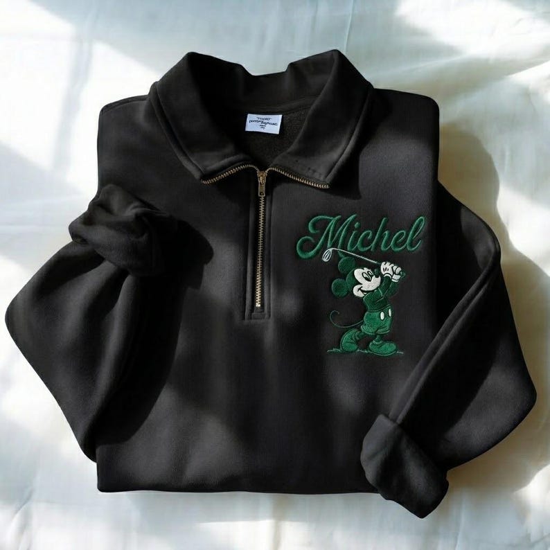 May include: Black sweatshirt with a collared neckline and a quarter-zip closure. The sweatshirt features an embroidered design of Mickey Mouse playing golf, with the name "Michel" in green script.