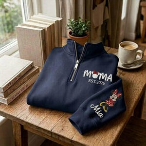 May include: Navy blue quarter-zip sweatshirt with "MOMA" and "EST.2026" in white script, featuring a Minnie Mouse design. The sleeve displays "Mia" and a Minnie Mouse graphic. The sweatshirt is on a wooden table.