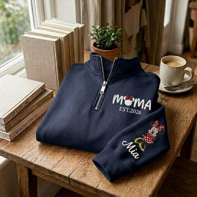 May include: Navy blue quarter-zip sweatshirt with "MOMA" and "EST.2026" in white script, featuring a Minnie Mouse design. The sleeve displays "Mia" and a Minnie Mouse graphic. The sweatshirt is on a wooden table.