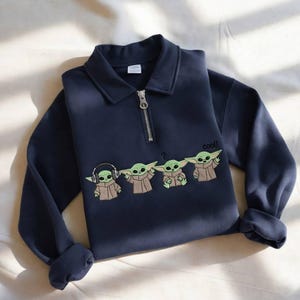 May include: Navy blue sweatshirt with a collared neckline and a quarter-zip closure. The sweatshirt features an embroidered design of four Grogu characters, with the word "cool!" next to them.