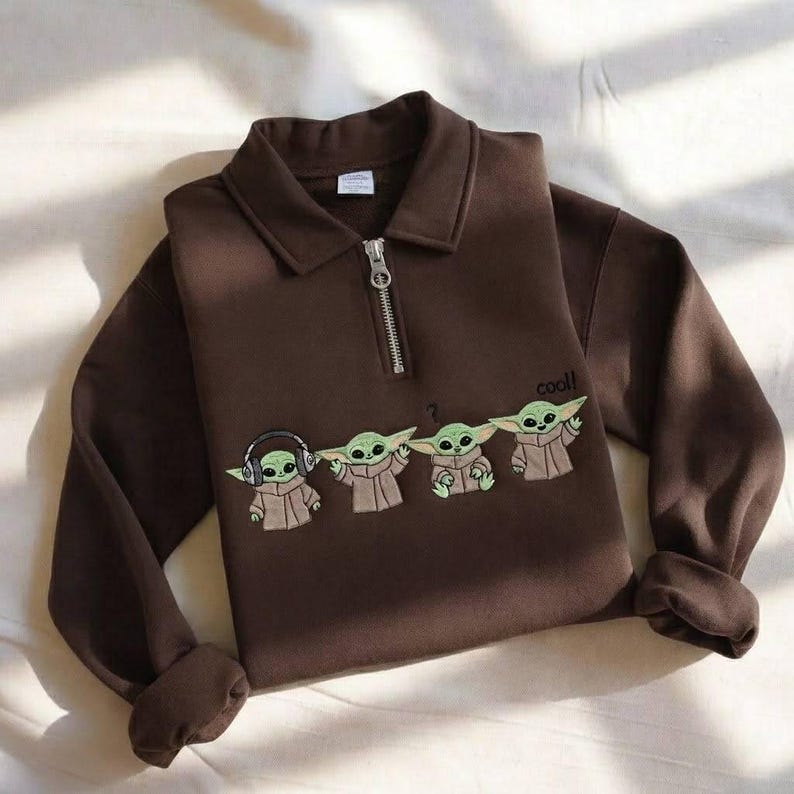 May include: A brown long-sleeve sweatshirt with a collared neckline and a silver zipper. The sweatshirt features an embroidered design of four green alien characters, with the word "cool!" next to them.