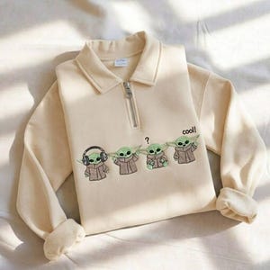 May include: Cream-colored sweatshirt with a collared neckline and a quarter-zip closure. The front features an embroidered design of four Grogu characters, with one wearing headphones and the word "cool!" printed on the side.