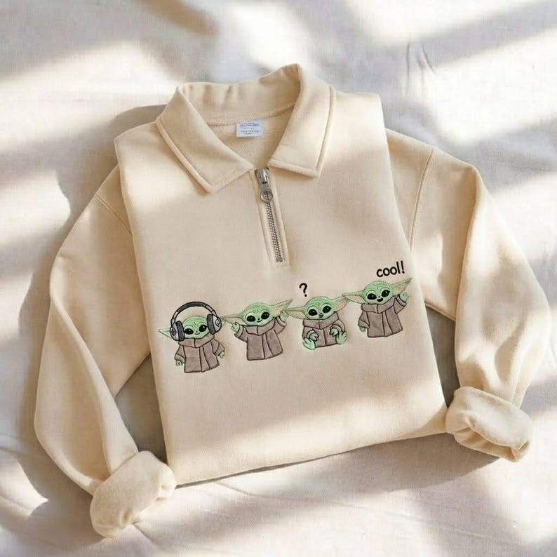May include: Cream-colored sweatshirt with a collared neckline and a quarter-zip closure. The front features an embroidered design of four Grogu characters, with one wearing headphones and the word "cool!" printed on the side.