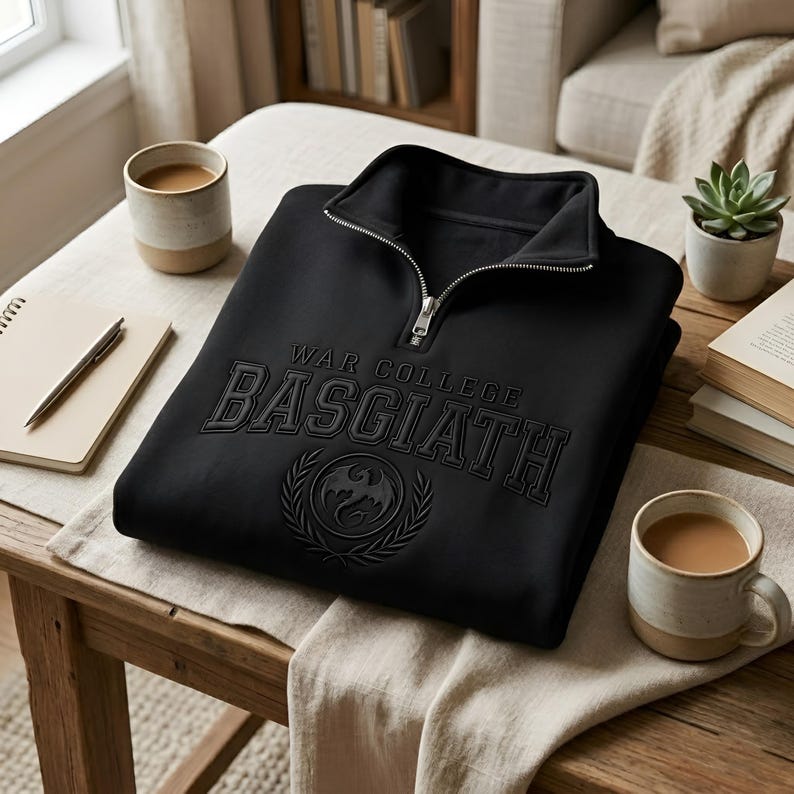 Puede incluir: Sudadera negra con cuello con cremallera. La sudadera presenta el texto "WAR COLLEGE BASGIATH" en relieve, junto con un emblema de drag&oacute;n. La sudadera est&aacute; doblada sobre una mesa de madera, con tazas de caf&eacute; y un cuaderno cerca.