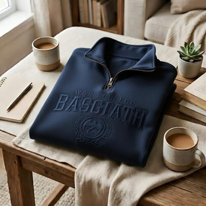 Puede incluir: Sudadera azul marino con cuello con cremallera. El texto "WAR COLLEGE BASCIATH" est&aacute; bordado en la parte delantera. La sudadera est&aacute; doblada sobre una mesa de madera. Otros objetos est&aacute;n alrededor de la mesa.