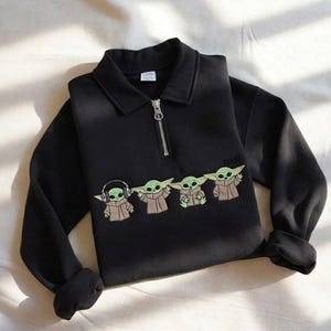 May include: Black sweatshirt with a collared neckline and a silver zipper. The sweatshirt features an embroidered design of four Grogu characters, with the word "cool" above the last character.