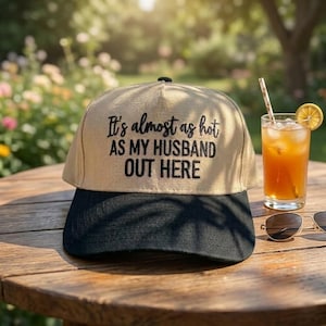 May include: A beige and black baseball cap with the text "It's almost as hot as my husband out here." The cap is on a wooden table next to a glass of iced tea with a lemon slice and sunglasses.