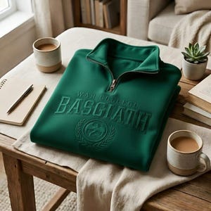 Puede incluir: Una sudadera verde esmeralda doblada con cuello con cremallera. La sudadera presenta el texto bordado "WAR COLLEGE BASCIATH" sobre un emblema circular. Tambi&eacute;n se ven dos tazas de caf&eacute;, un bloc de notas y un bol&iacute;grafo.