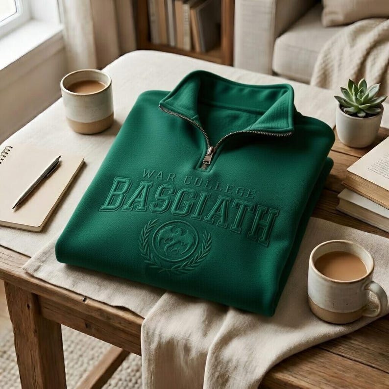 Puede incluir: Una sudadera verde esmeralda doblada con cuello con cremallera. La sudadera presenta el texto bordado "WAR COLLEGE BASCIATH" sobre un emblema circular. Tambi&eacute;n se ven dos tazas de caf&eacute;, un bloc de notas y un bol&iacute;grafo.