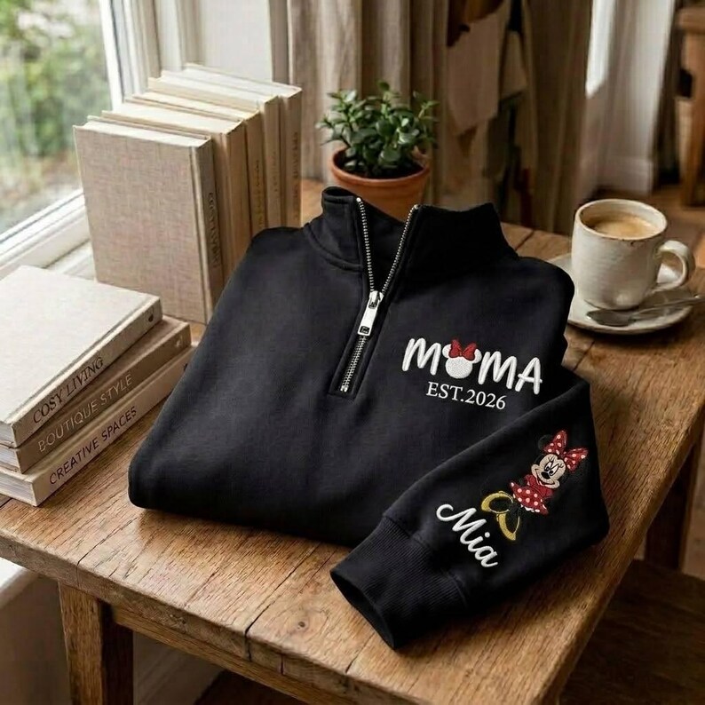 May include: Black quarter-zip sweatshirt with white "MOMA EST. 2026" text and a Minnie Mouse graphic. The sleeve features the name "Mia" and a Minnie Mouse design. The sweatshirt is displayed on a wooden table.