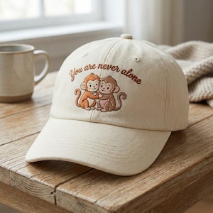 Peut inclure: Casquette de baseball crème avec une visière incurvée. Le chapeau présente un motif brodé de deux singes enlacés et le texte "You are never alone" en script marron. Le chapeau est posé sur une surface en bois.
